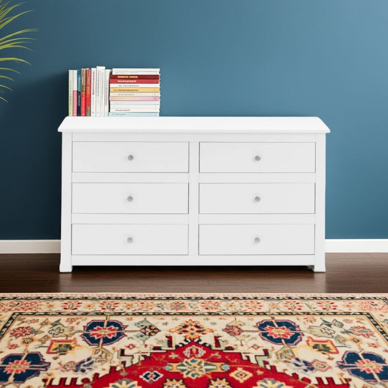 White dresser with 6 drawers and books against a blue wall with a patterned rug on the floor.