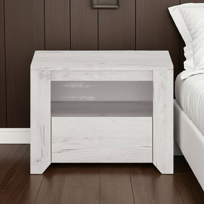 White wooden nightstand against a dark wood paneled wall with a bed in the background.