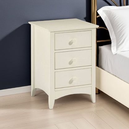 White nightstand with three drawers next to a bed in a bedroom setting.