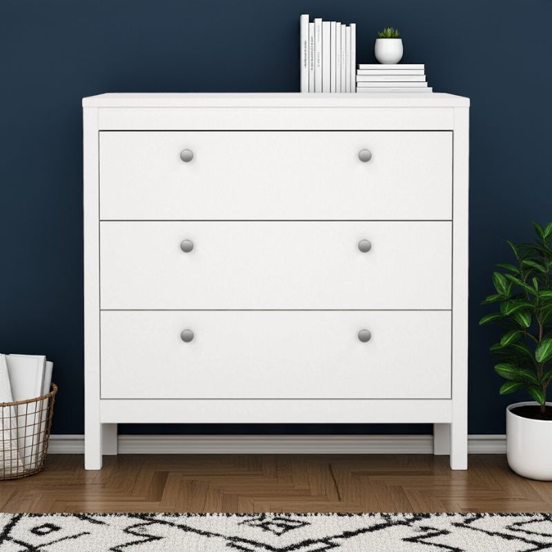 White dresser with three drawers against a dark blue wall, with books and a plant on top.