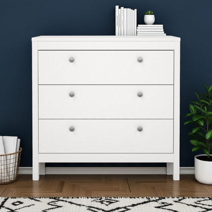 White dresser with three drawers against a dark blue wall, with books and a plant on top.