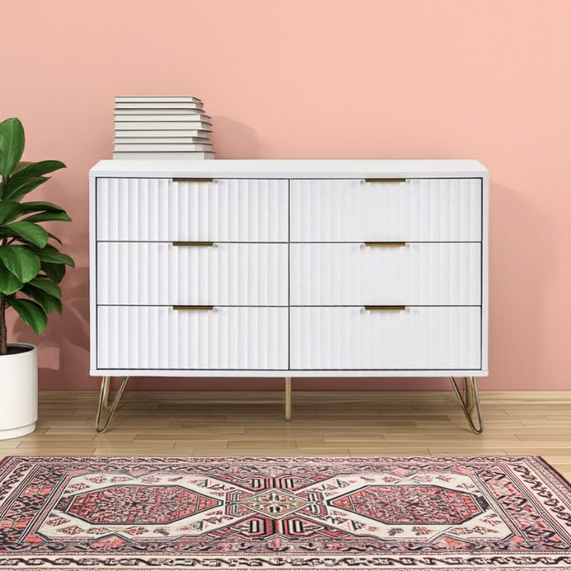 White dresser with gold handles against a pink wall with a plant and rug.