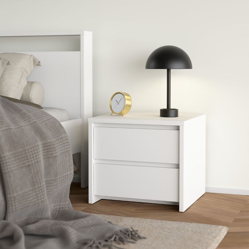 White nightstand with black lamp and gold clock next to a bed in a bedroom.