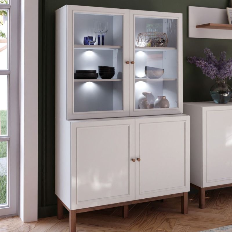 White display cabinet with glass doors in a room with a window and another piece of furniture.