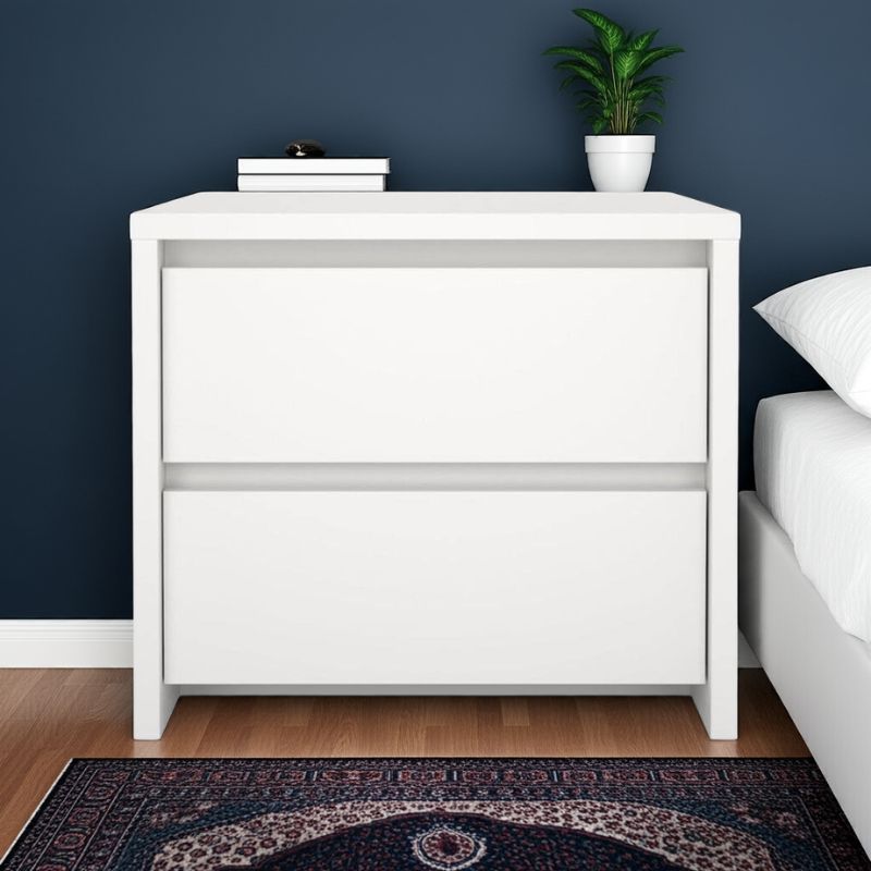 White bedside table with books and a plant against a dark blue wall.